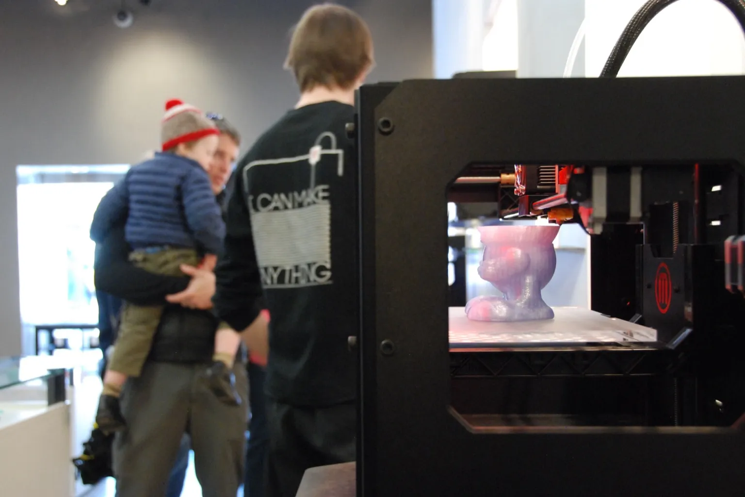 A black MakerBot 3D printer on display in a store, printing a pink and purple figure, with shoppers behind it: a man in a shirt printed "I can make anything" and a man carrying a small child