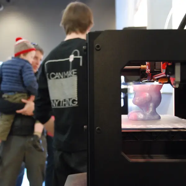 A black MakerBot 3D printer on display in a store, printing a pink and purple figure, with shoppers behind it: a man in a shirt printed "I can make anything" and a man carrying a small child