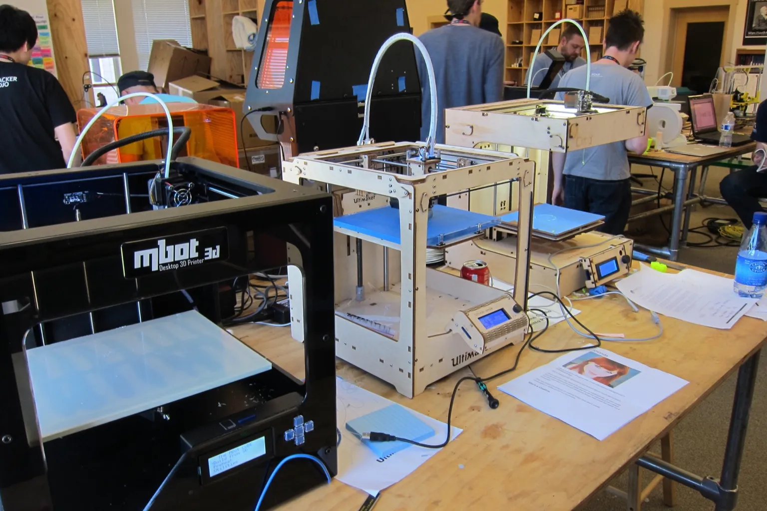 Three desktop 3D printers side by side on a long table in a busy workshop: a black mbot 3D, a plywood Ultimaker and a second plywood-framed printer, with more printers and people behind them