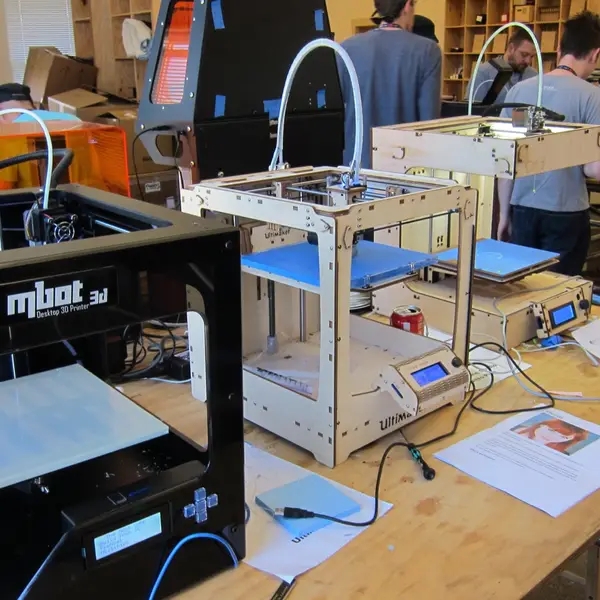 Three desktop 3D printers side by side on a long table in a busy workshop: a black mbot 3D, a plywood Ultimaker and a second plywood-framed printer, with more printers and people behind them