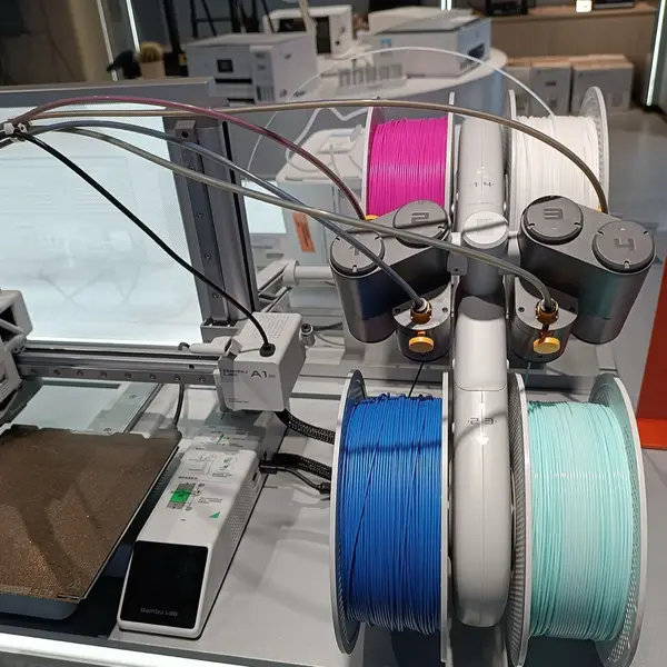 A Bambu Lab A1 mini on a store display table beside an AMS lite with feeders numbered 1 to 4, holding pink, white, blue and mint-green spools, its four PTFE tubes and cable clipped together and running to the printer's toolhead
