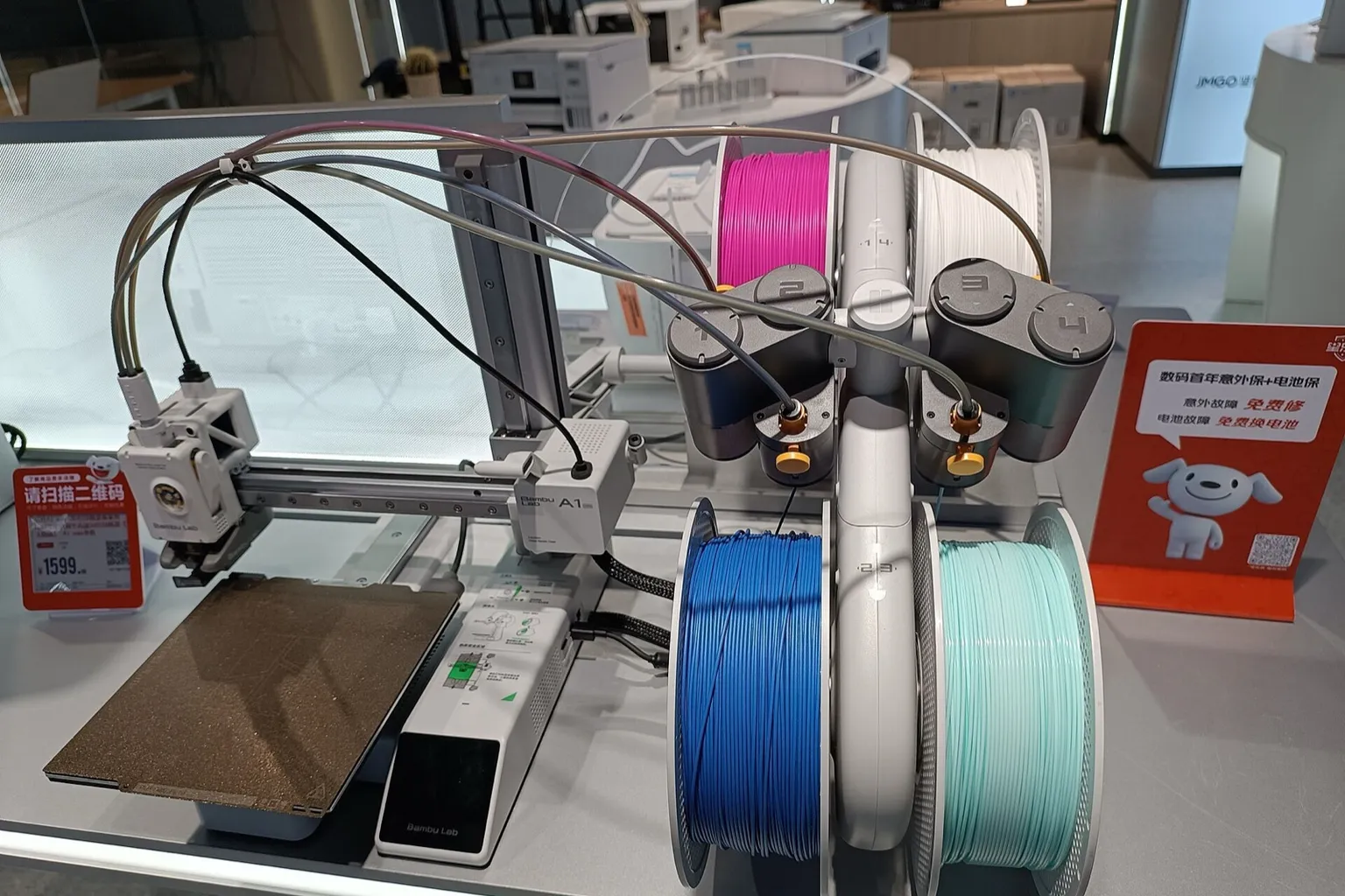 A Bambu Lab A1 mini on a store display table beside an AMS lite with feeders numbered 1 to 4, holding pink, white, blue and mint-green spools, its four PTFE tubes and cable clipped together and running to the printer's toolhead