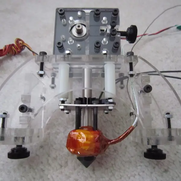 A hand-built 3D printer extruder with clear acrylic plates, a drive gear, black thumb screws and a hotend wrapped in orange Kapton tape, lying on a carpet