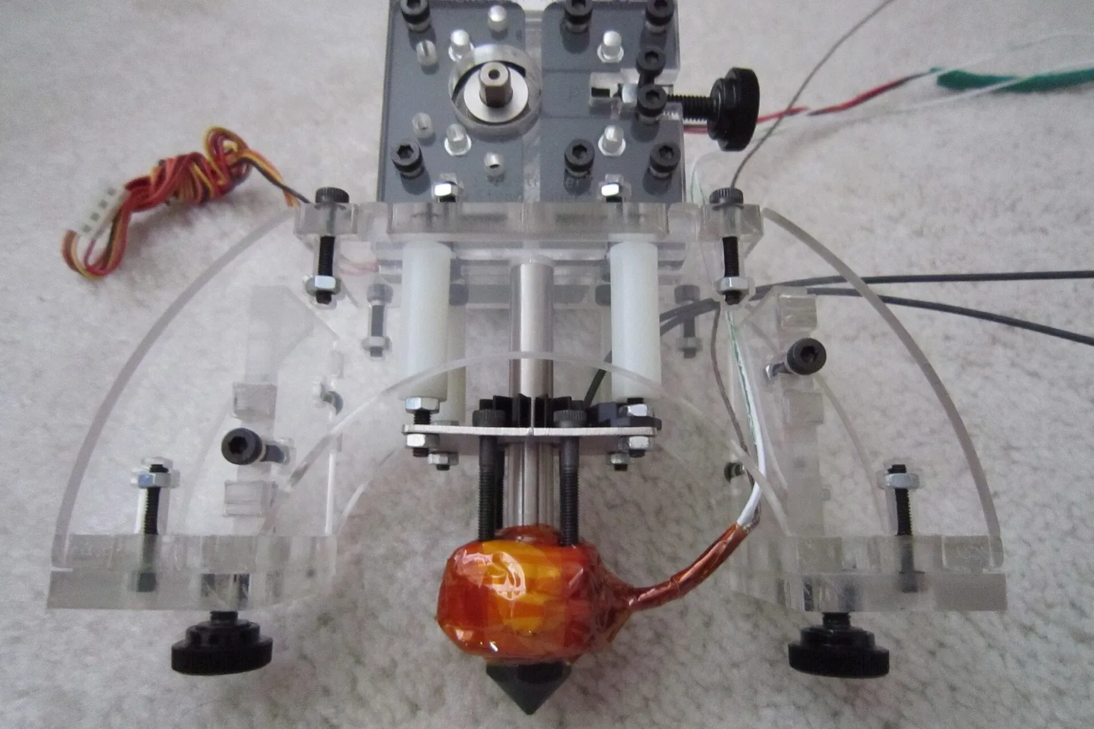 A hand-built 3D printer extruder with clear acrylic plates, a drive gear, black thumb screws and a hotend wrapped in orange Kapton tape, lying on a carpet