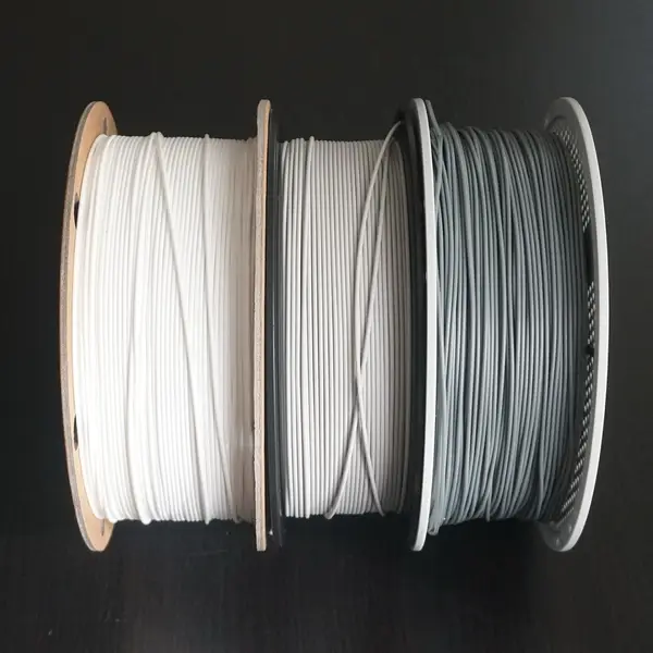 Three filament spools side by side on a dark table: white filament on a cardboard spool, light gray filament on a black plastic spool and gray refill filament on a white reusable spool