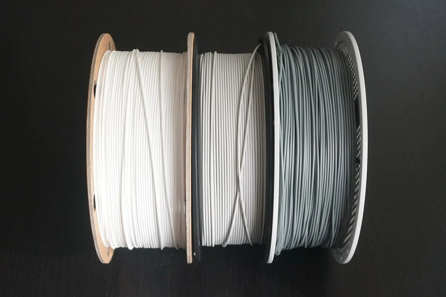 Three filament spools side by side on a dark table: white filament on a cardboard spool, light gray filament on a black plastic spool and gray refill filament on a white reusable spool