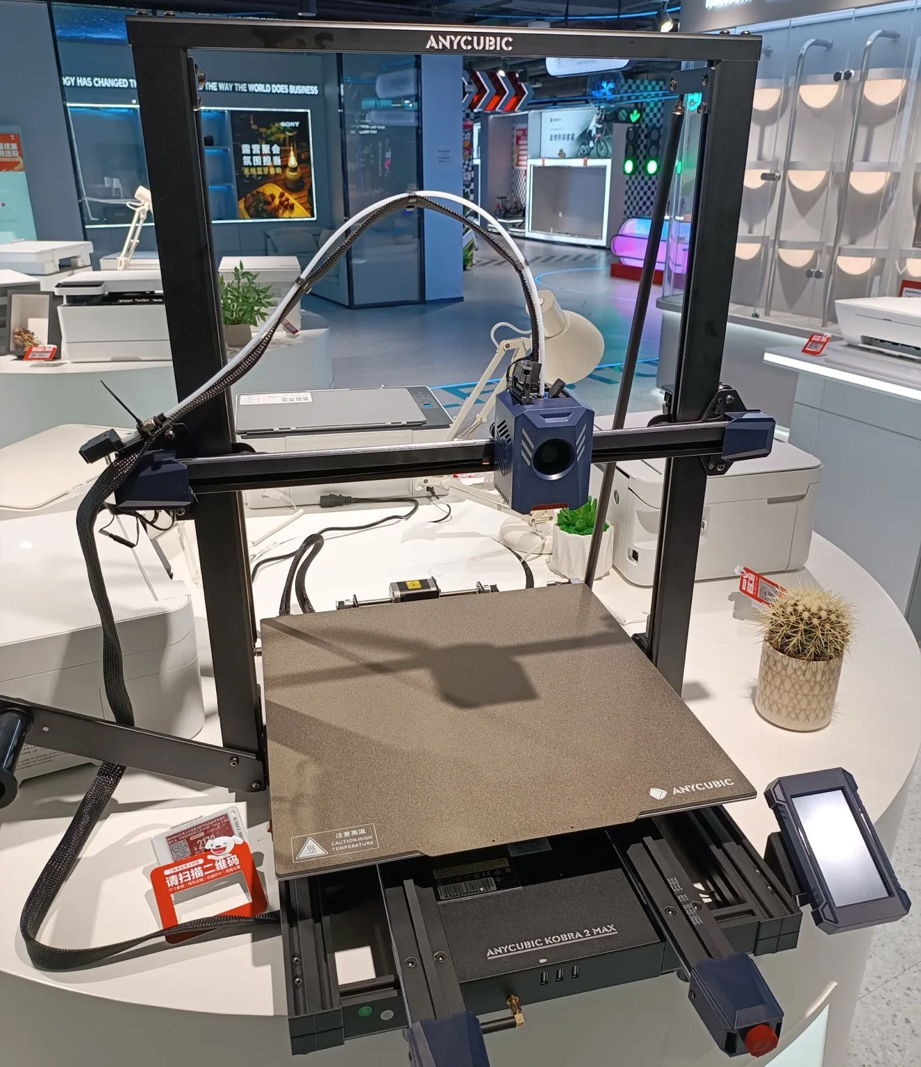 An Anycubic Kobra 2 Max FDM printer on a white display table in an electronics store, with its blue print head on the gantry above a textured build plate