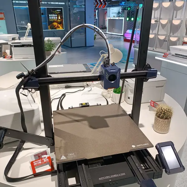 An Anycubic Kobra 2 Max FDM printer on a white display table in an electronics store, with its blue print head on the gantry above a textured build plate