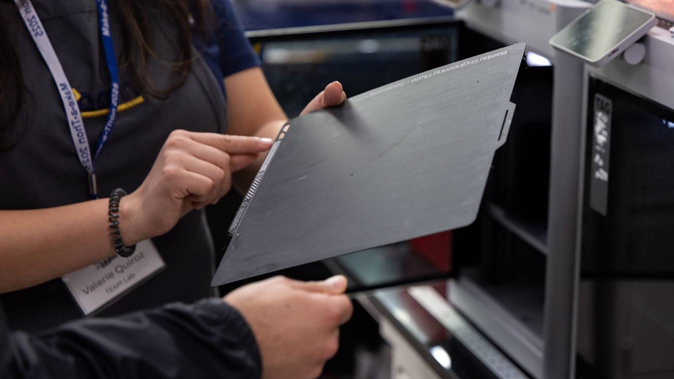 A person holding a dark gray Bambu Engineering Plate, its name printed along the top edge, in front of a row of Bambu Lab printers