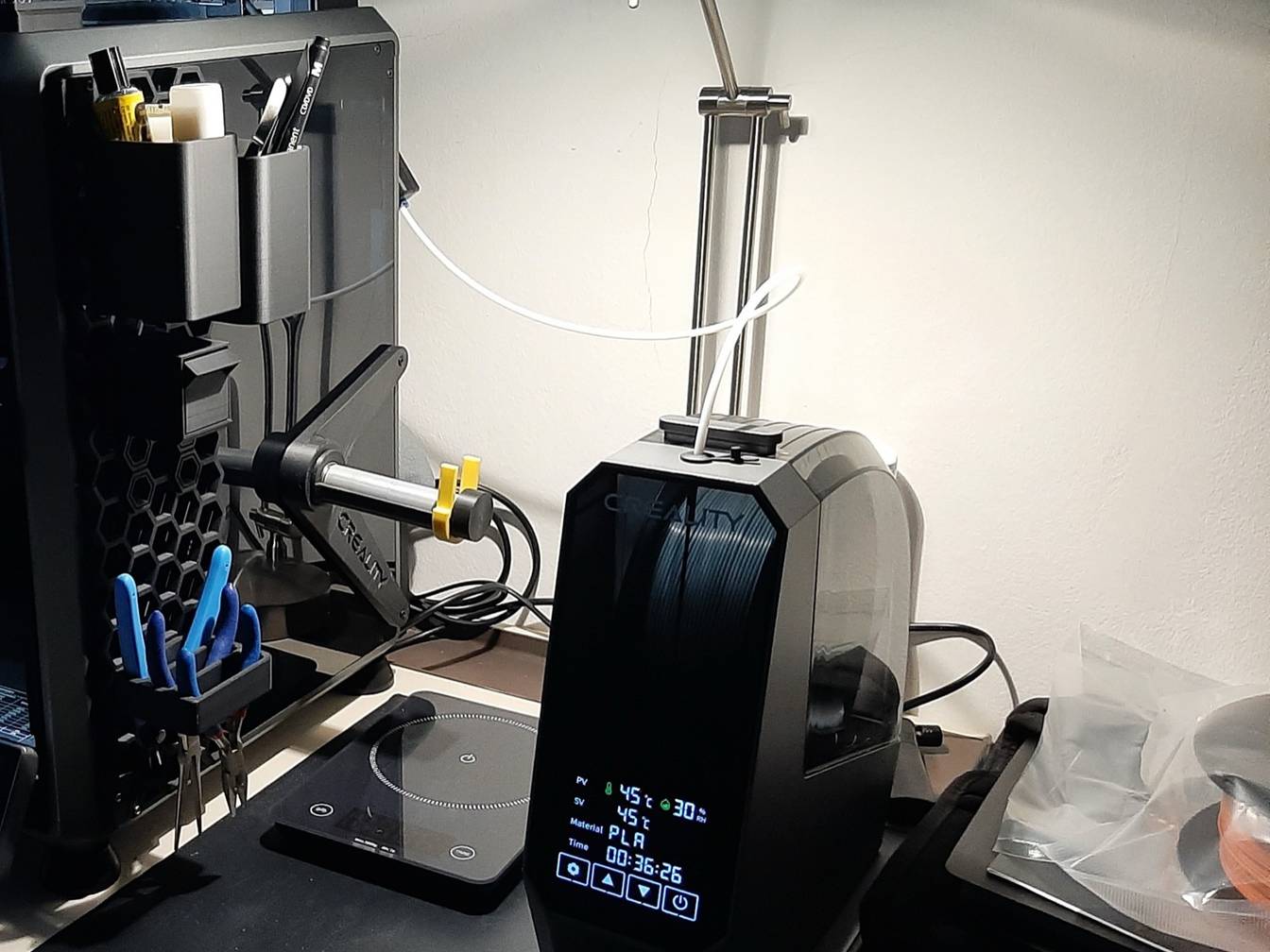 A black filament dryer holding a spool, its display reading 45 °C, PLA and a running timer, with filament fed out through a white tube, next to the side of an enclosed 3D printer