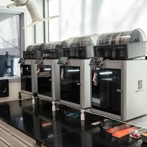 Four Bambu Lab X1 Carbon printers in a row on a black lab bench by a window, each with an AMS unit on top and its touchscreen lit
