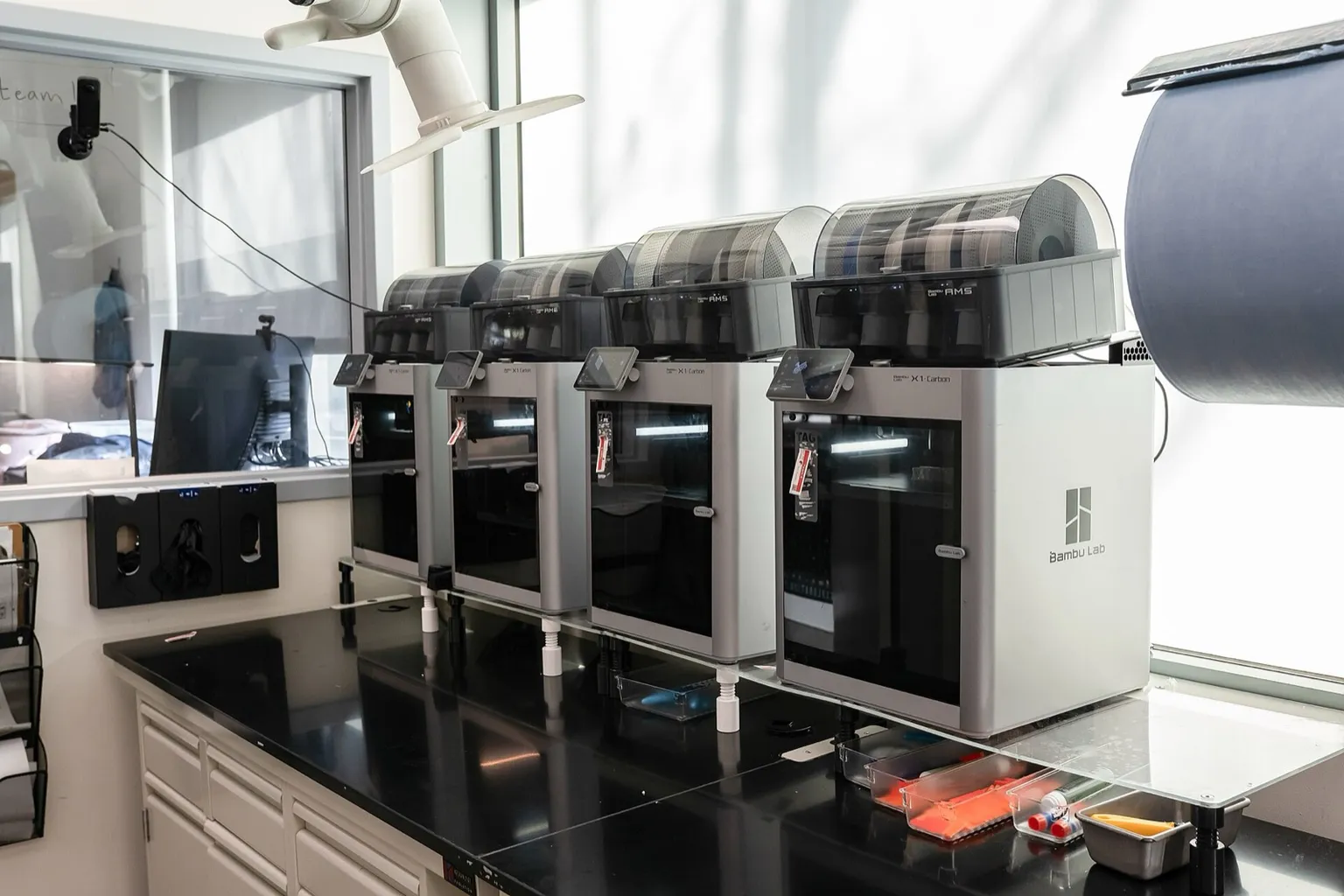 Four Bambu Lab X1 Carbon printers in a row on a black lab bench by a window, each with an AMS unit on top and its touchscreen lit