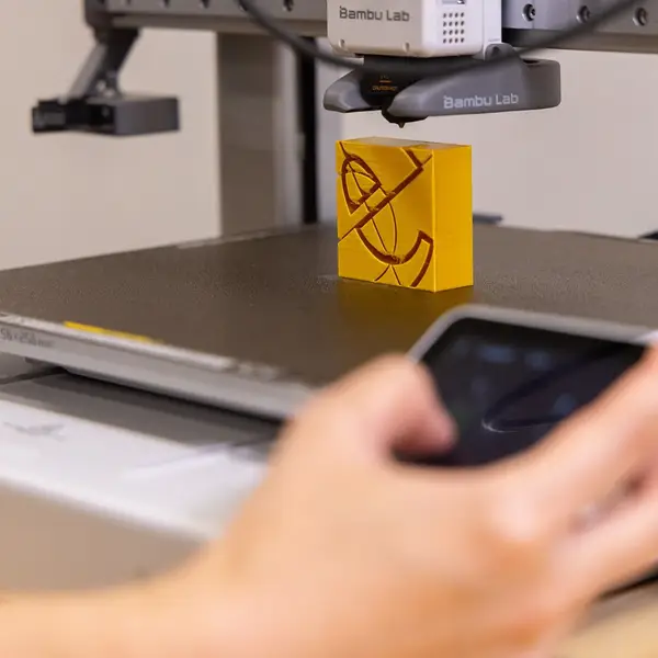 A hand reaches for the touchscreen of a Bambu Lab A1, blurred in the foreground, below the toolhead and a yellow printed block on the build plate