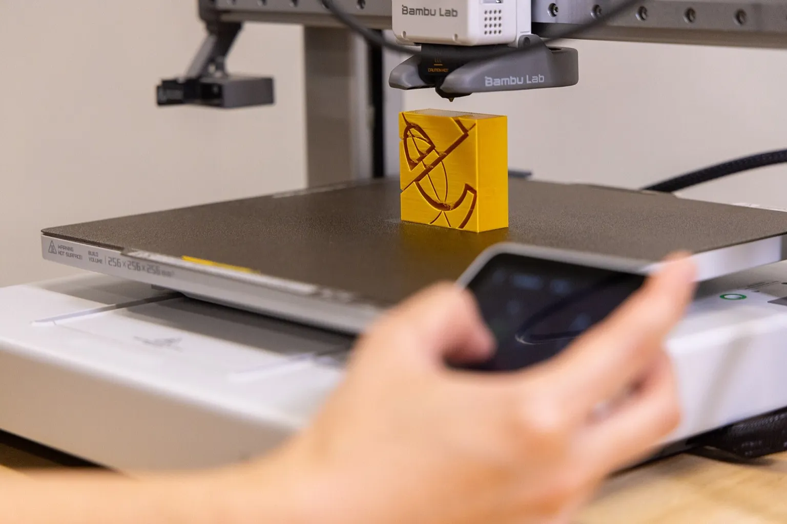 A hand reaches for the touchscreen of a Bambu Lab A1, blurred in the foreground, below the toolhead and a yellow printed block on the build plate