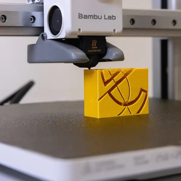 Close-up of a Bambu Lab A1 toolhead, its nozzle under a black silicone sock marked Caution Hot, printing a yellow part on a textured build plate