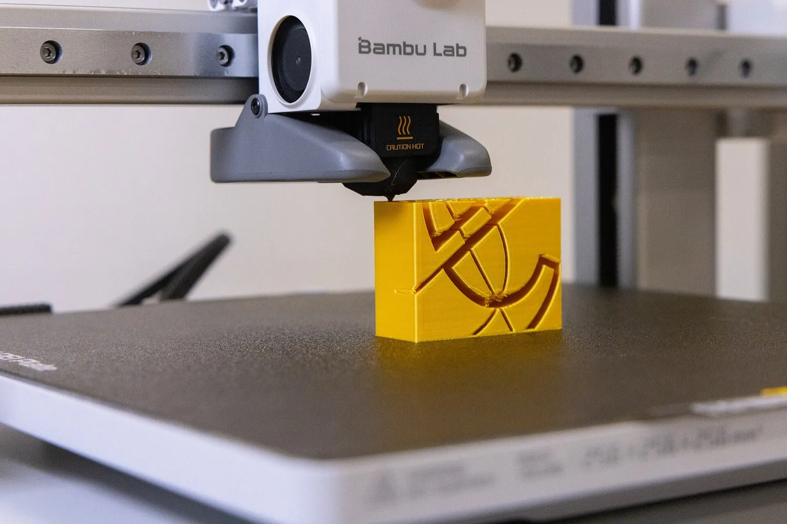 Close-up of a Bambu Lab A1 toolhead, its nozzle under a black silicone sock marked Caution Hot, printing a yellow part on a textured build plate