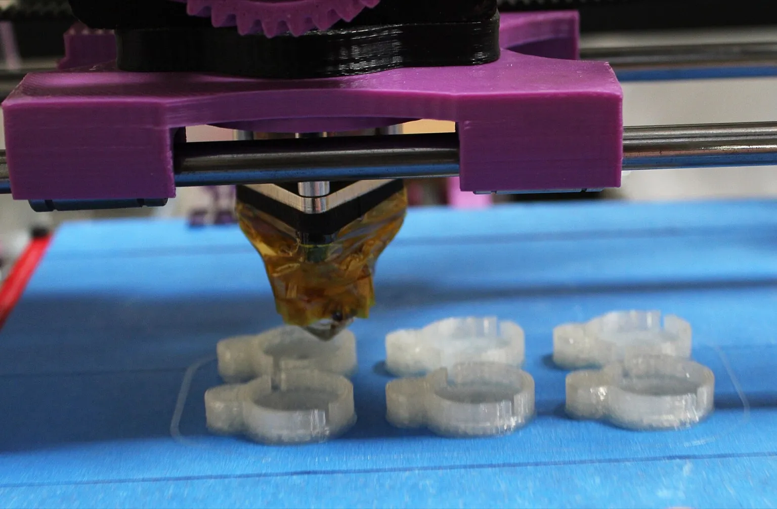 A hotend wrapped in amber Kapton tape hangs from a purple printed carriage on steel rods, printing six small clear parts on a blue-taped bed