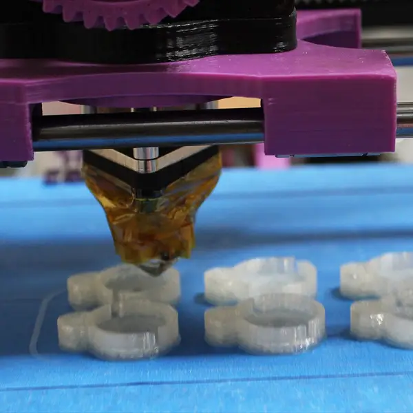 A hotend wrapped in amber Kapton tape hangs from a purple printed carriage on steel rods, printing six small clear parts on a blue-taped bed