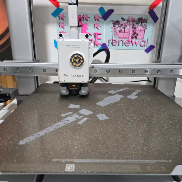 A Bambu Lab A1 toolhead over a textured build plate, its white cover printed with "Remove this cover for hotend replacement", with a first layer of grey parts on the plate
