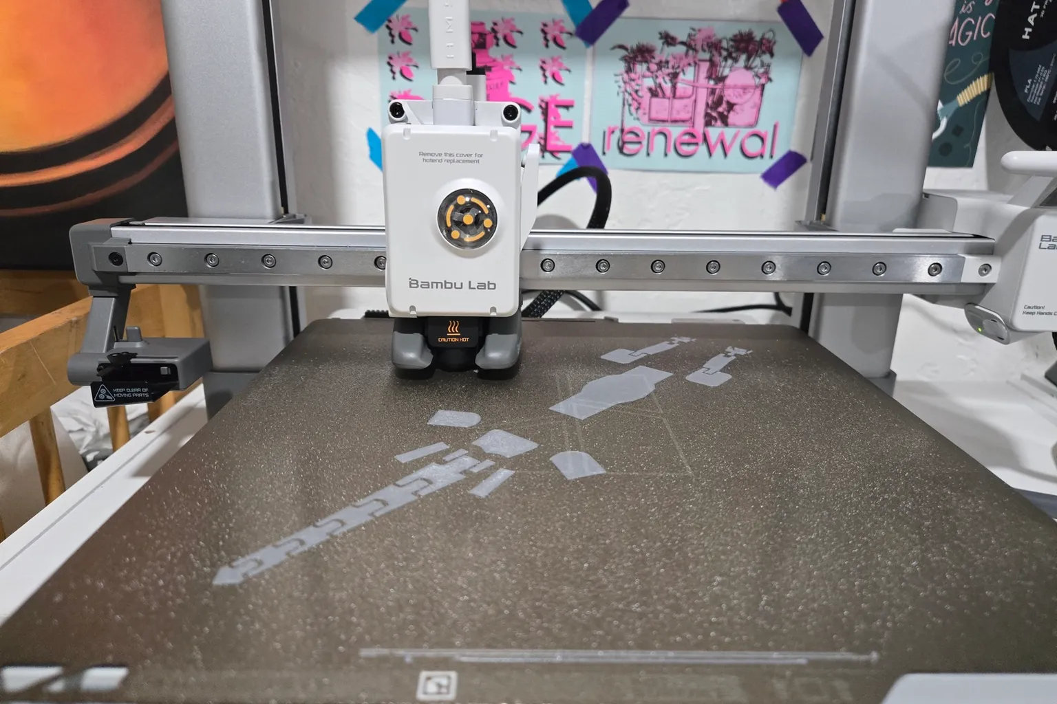 A Bambu Lab A1 toolhead over a textured build plate, its white cover printed with "Remove this cover for hotend replacement", with a first layer of grey parts on the plate