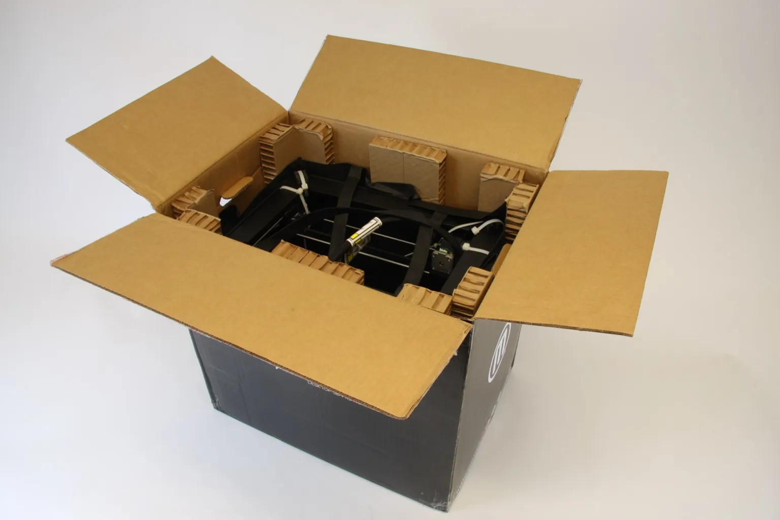 An opened cardboard shipping box, seen from above, with honeycomb cardboard blocks holding the strapped black frame of a MakerBot Replicator 2 3D printer inside