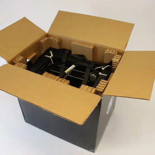 An opened cardboard shipping box, seen from above, with honeycomb cardboard blocks holding the strapped black frame of a MakerBot Replicator 2 3D printer inside