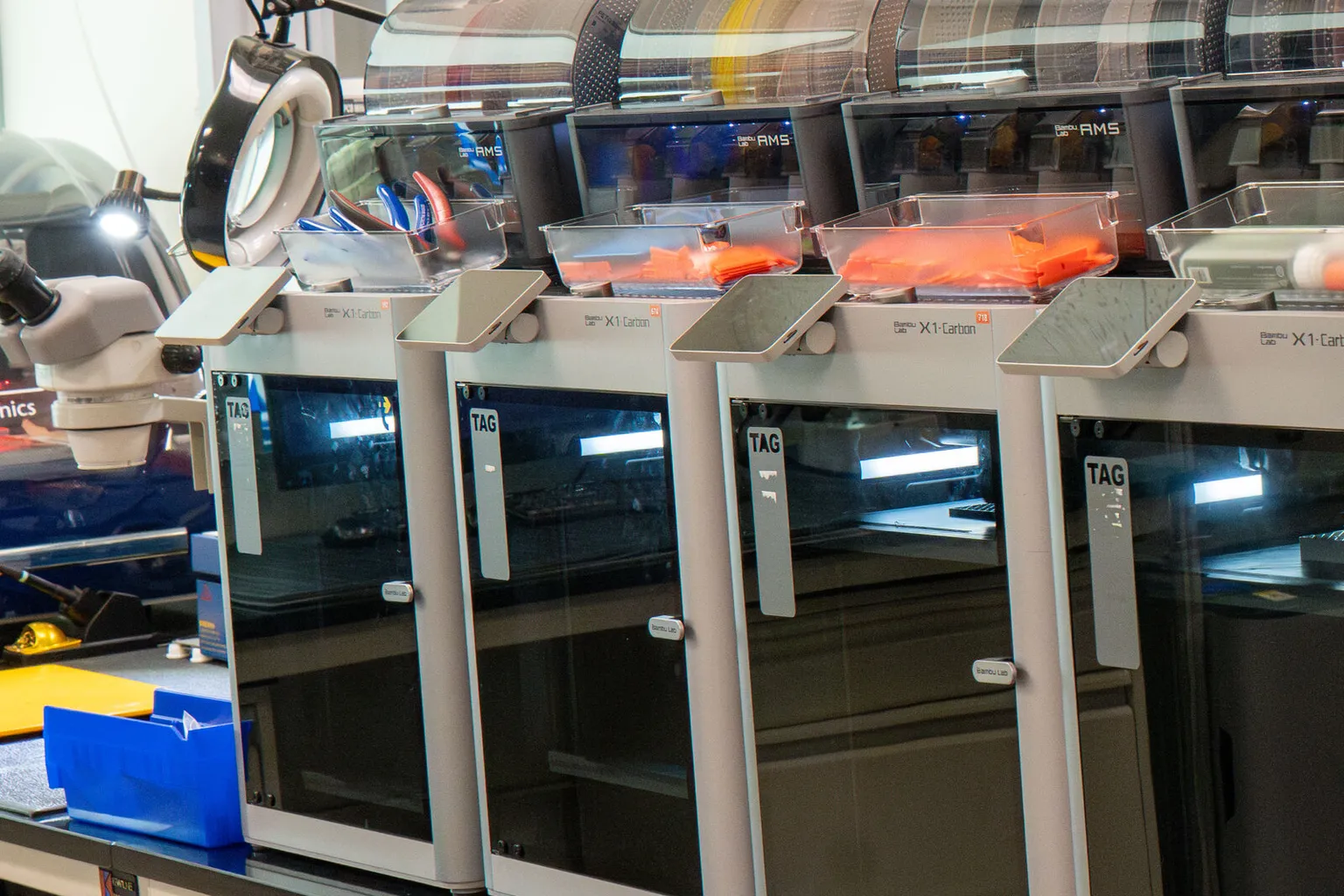 A row of Bambu Lab X1 Carbon printers on a lab bench, each with an AMS unit and a clear parts tray on top