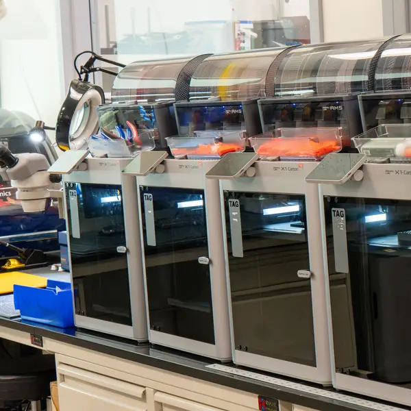 A row of Bambu Lab X1 Carbon printers on a lab bench, each with an AMS unit and a clear parts tray on top