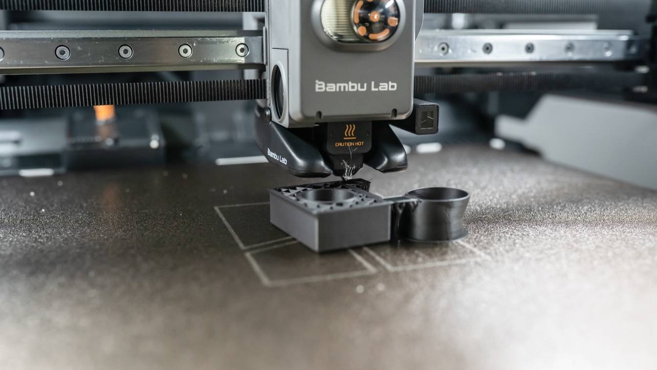 A Bambu Lab toolhead printing a dark gray part on a speckled bronze textured build plate, with faint rectangular marks on the plate around the part