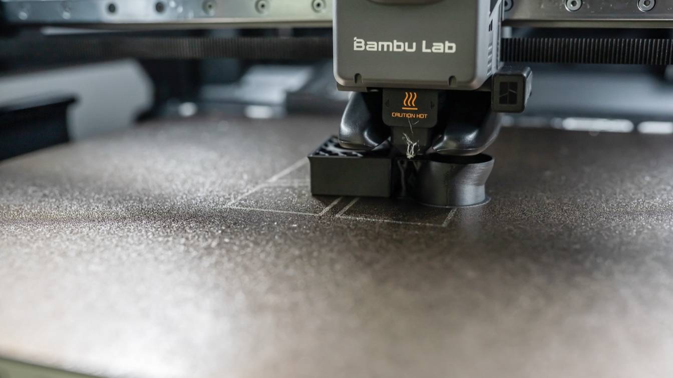 Close-up of a Bambu Lab X1 Carbon toolhead printing a black part on a textured build plate, with a wisp of filament at the nozzle