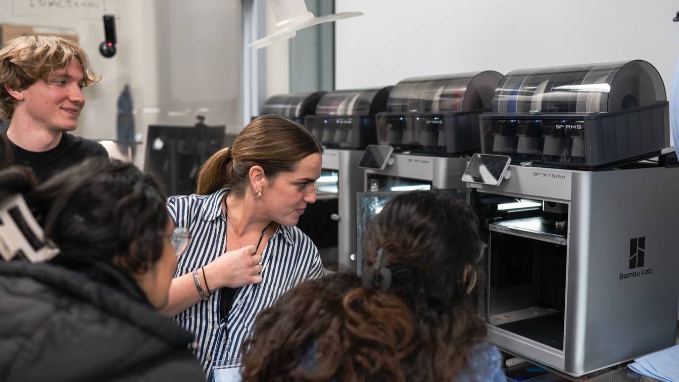 A row of enclosed Bambu Lab X1 Carbon printers with AMS units on top, lined up on a lab bench, with students gathered in front of them