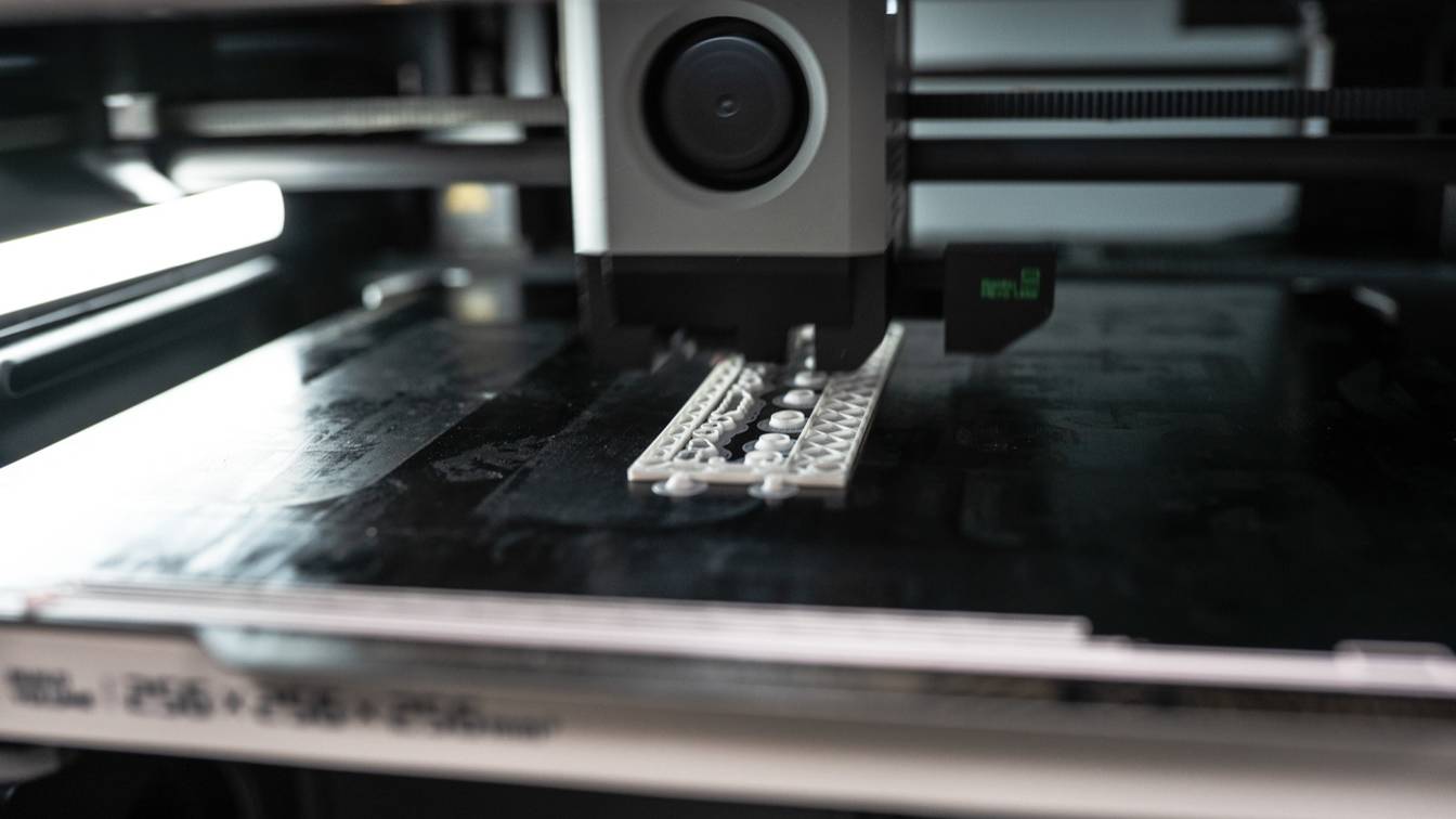 Close-up inside an enclosed Bambu Lab printer: the toolhead lays down a white lattice part on a dark build plate, lit by the chamber light