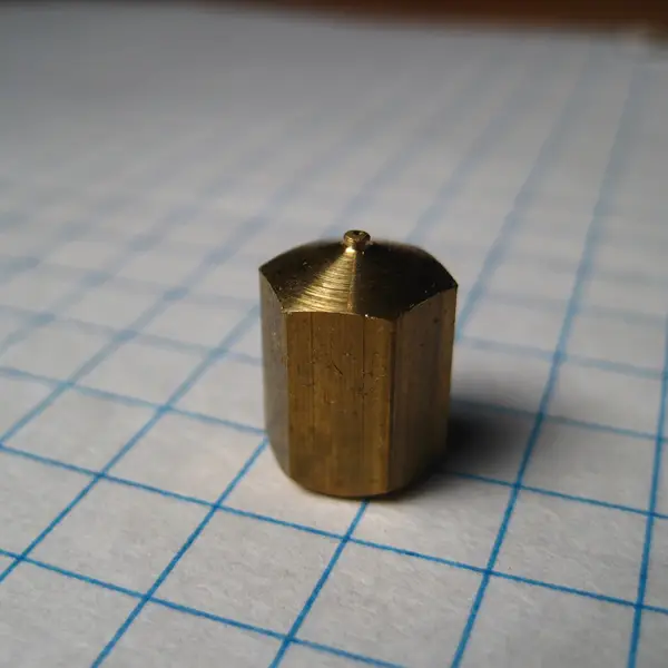 A machined brass hexagonal extruder nozzle for a RepRap printer standing on blue-ruled graph paper