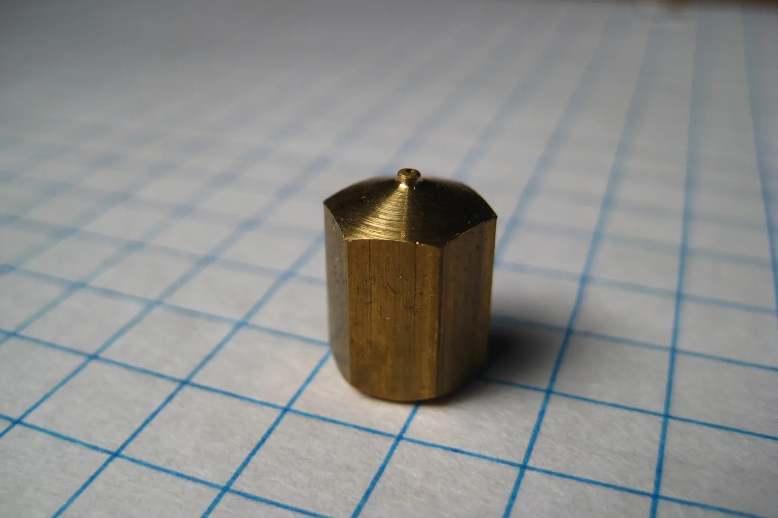 A machined brass hexagonal extruder nozzle for a RepRap printer standing on blue-ruled graph paper