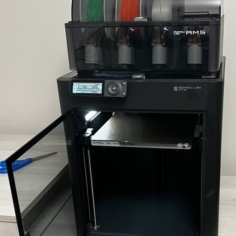 A Bambu Lab P1S with its glass front door open and a Bambu Lab AMS on top holding green, orange and white filament spools
