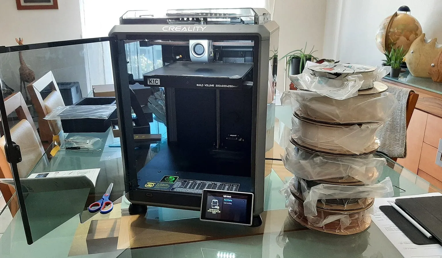 An enclosed Creality K1C printer on a desk beside a stack of filament spools