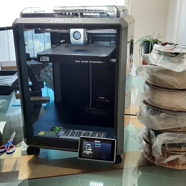 An enclosed Creality K1C printer on a desk beside a stack of filament spools