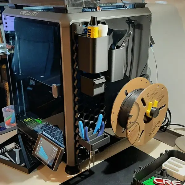 A dark grey Creality K1C 3D printer on a workbench, with printed tool holders, pliers and a filament spool mounted on its side