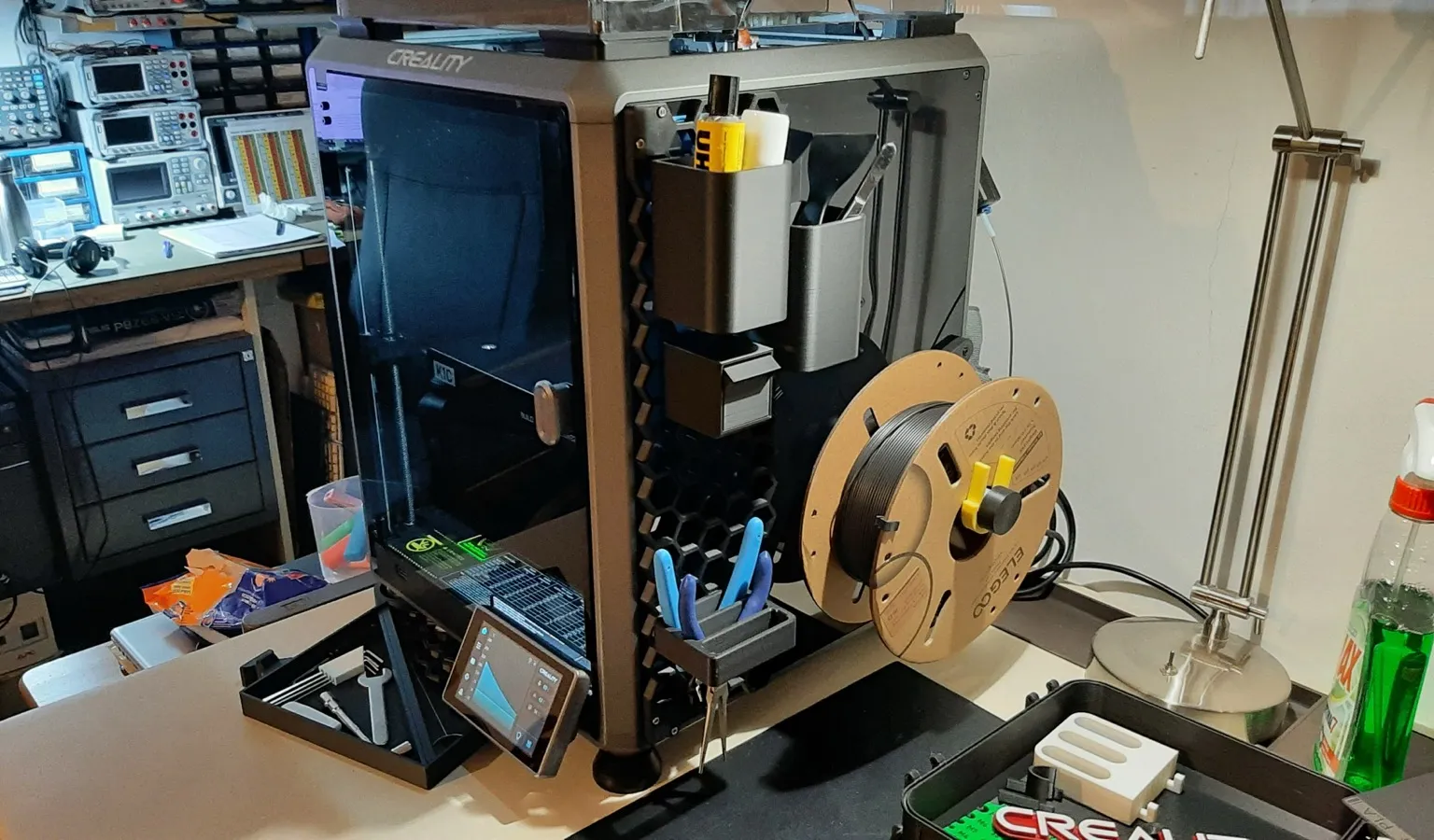 A dark grey Creality K1C 3D printer on a workbench, with printed tool holders, pliers and a filament spool mounted on its side