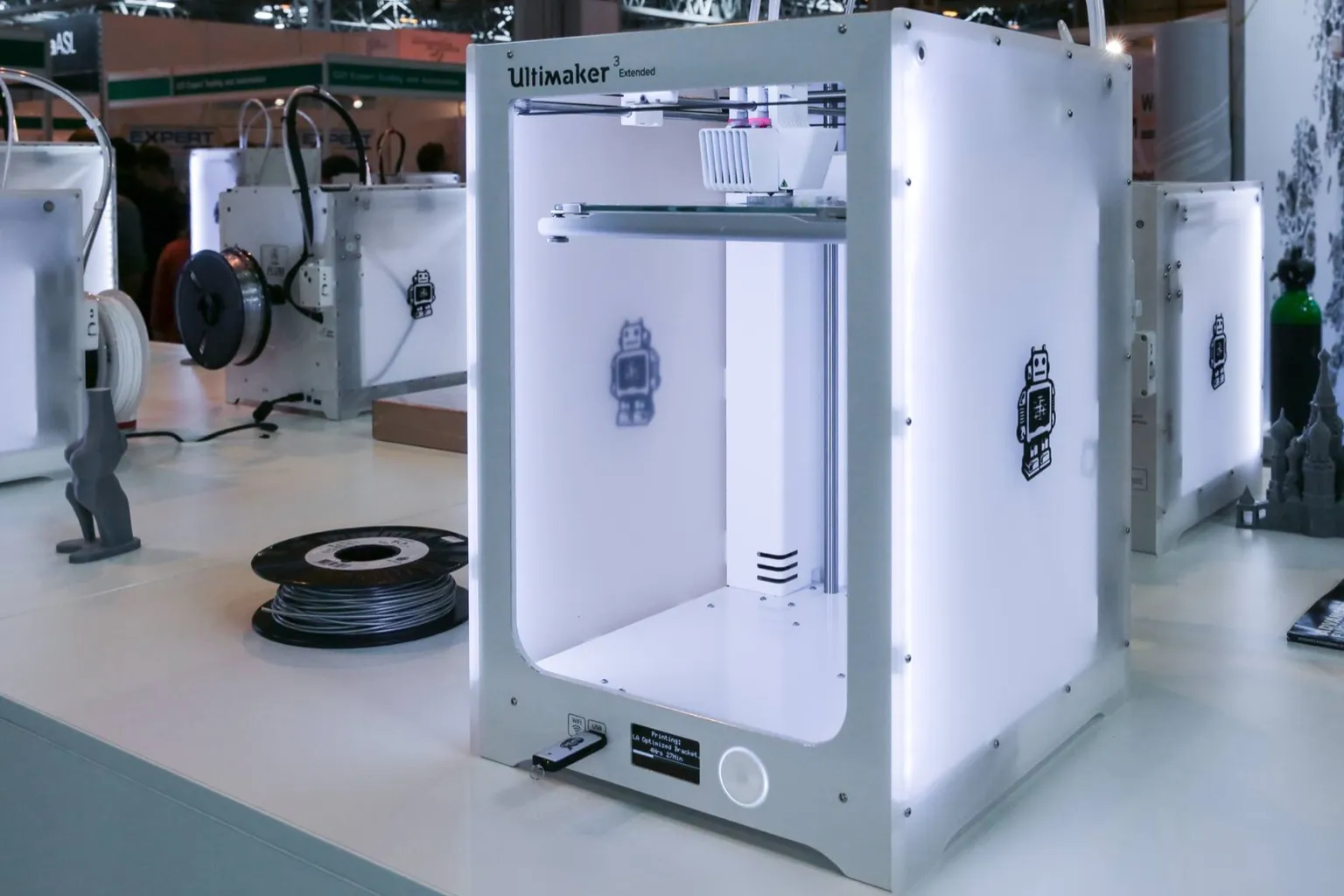 A white UltiMaker 3 Extended printer with translucent side panels on an exhibition stand, with a spool of grey filament beside it