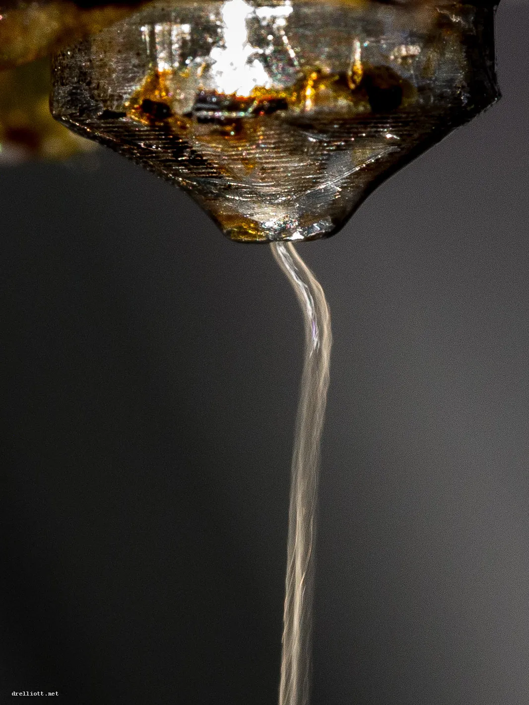 Close-up of a 3D printer nozzle with a thin strand of molten plastic hanging from its tip