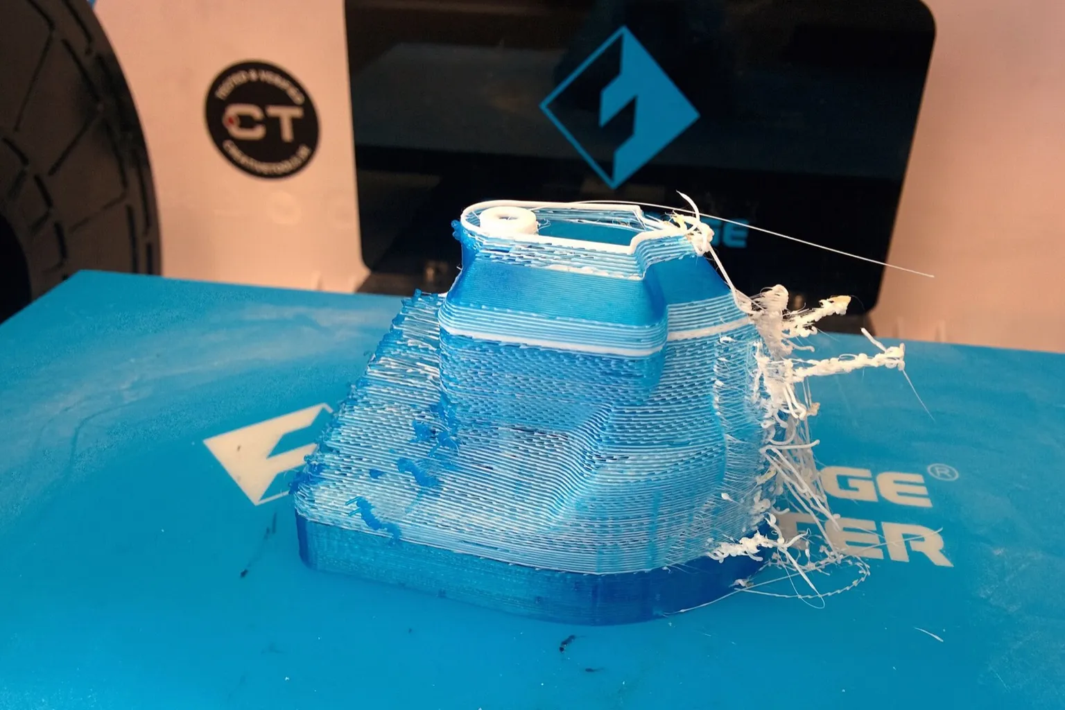 A half-printed blue and white 3DBenchy with white strings trailing off one side, on the blue build plate of a dual-extrusion printer