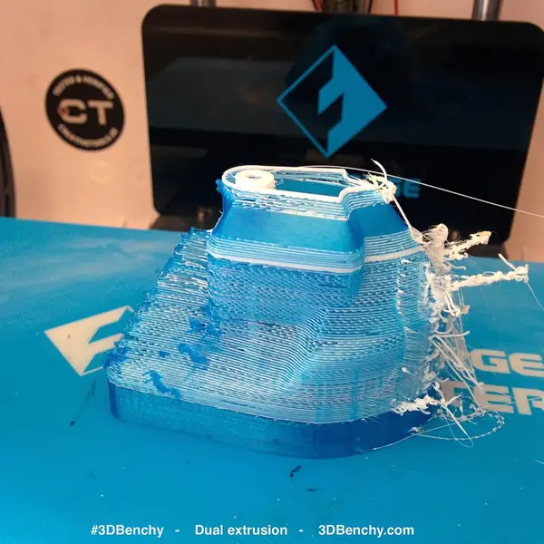 A half-printed blue and white 3DBenchy with white strings trailing off one side, on the blue build plate of a dual-extrusion printer