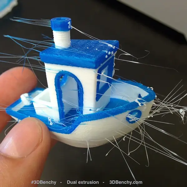 A hand holding a blue and white 3DBenchy boat printed with dual extrusion, with thin strings of plastic trailing off it