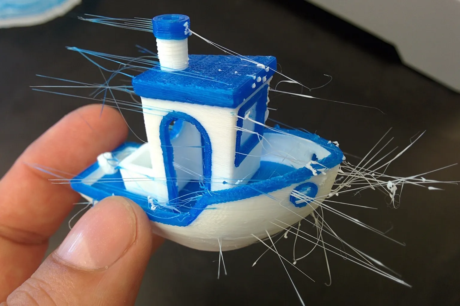 A hand holding a blue and white 3DBenchy boat printed with dual extrusion, with thin strings of plastic trailing off it