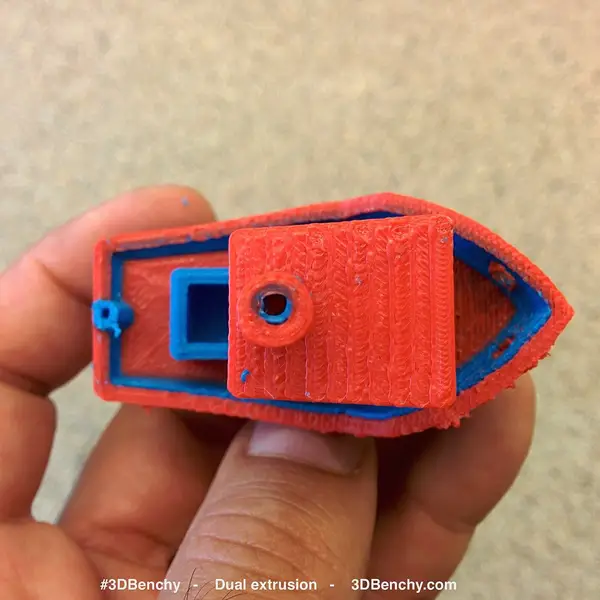 A hand holding a small boat model printed in red and blue plastic, seen from above, with the 3DBenchy name printed along the bottom edge