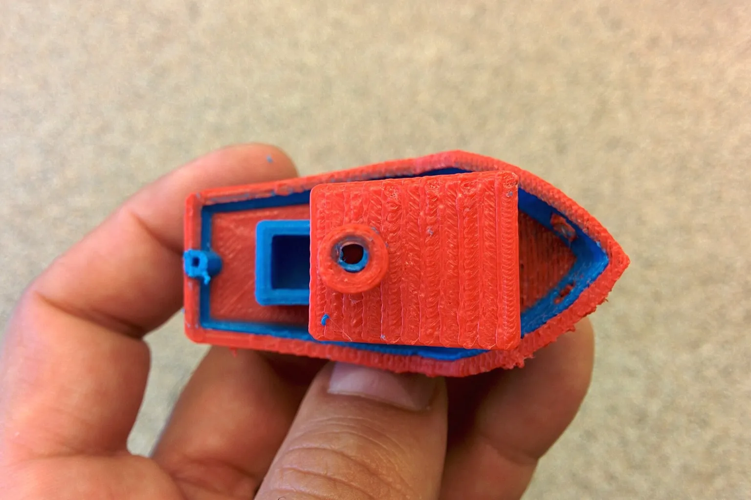 A hand holding a small boat model printed in red and blue plastic, seen from above, with the 3DBenchy name printed along the bottom edge