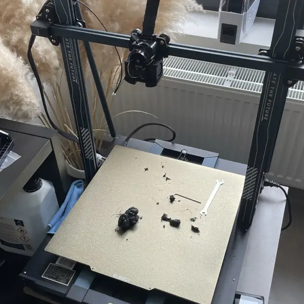 An Elegoo Neptune 4 Max FDM printer seen from a high angle, with blobs of black filament, a hex key and a wrench on its gold textured build plate