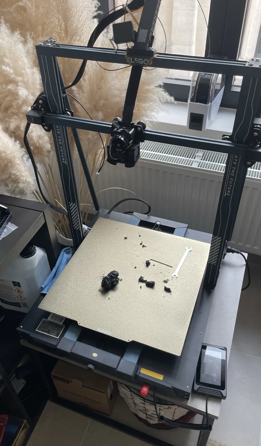 An Elegoo Neptune 4 Max FDM printer seen from a high angle, with blobs of black filament, a hex key and a wrench on its gold textured build plate