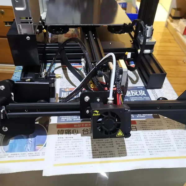 The X gantry of a Creality Ender 3 during kit assembly, with the stock Bowden extruder at its left end and a white PTFE tube rising from the hotend under the fan shroud on the carriage
